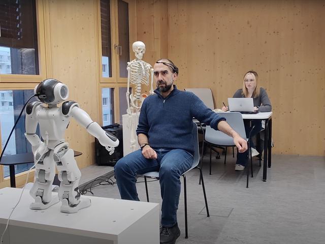The image shows a humanoid robot interacting with a person wearing VR goggles in an indoor setting.