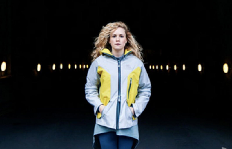 female standing in dark tunnel, wearing a light grey waterproof jacket with yellow patches on sides