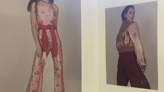 two exhibit pictures of two female models. Left model is wearing a pink floral print wide leg jumpsuit that is tied at the waist with a dark pink sash and the left model is wearing a metallic bronze bomber jacket with pink sleeves and a heart print, with brown wide leg trousers 
