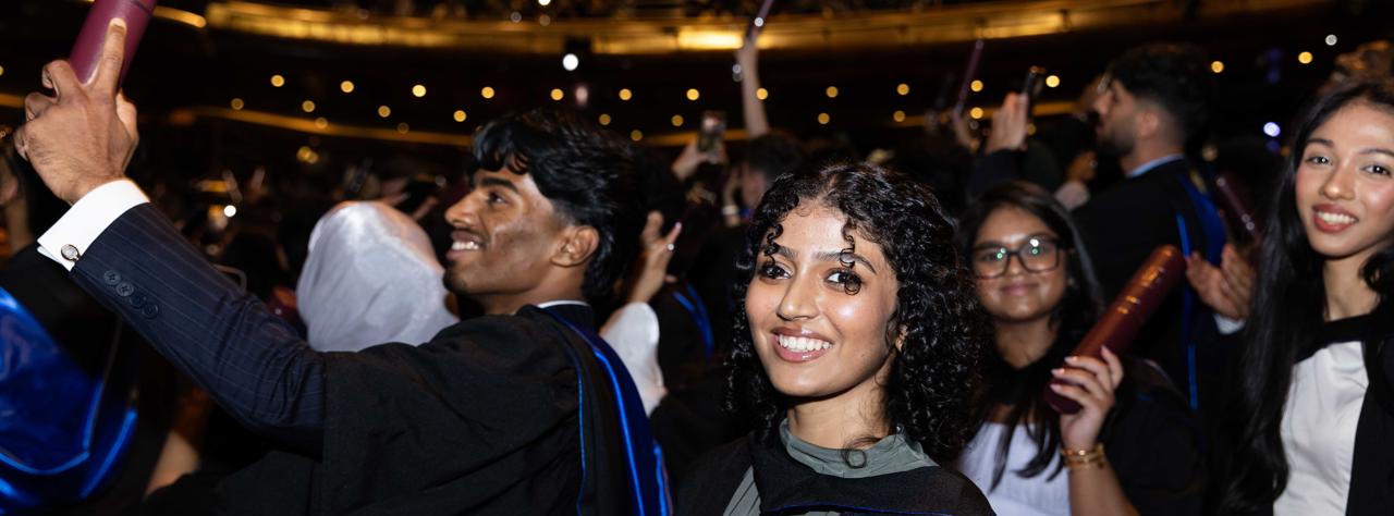 student wearing a graduation gown and holding a scroll looking at camera with other students cheering in the background