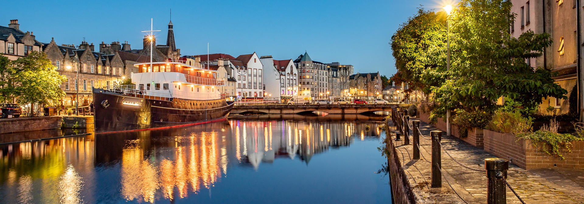 A city river at dusk, featuring a boat on the water, surrounded by evening lights.