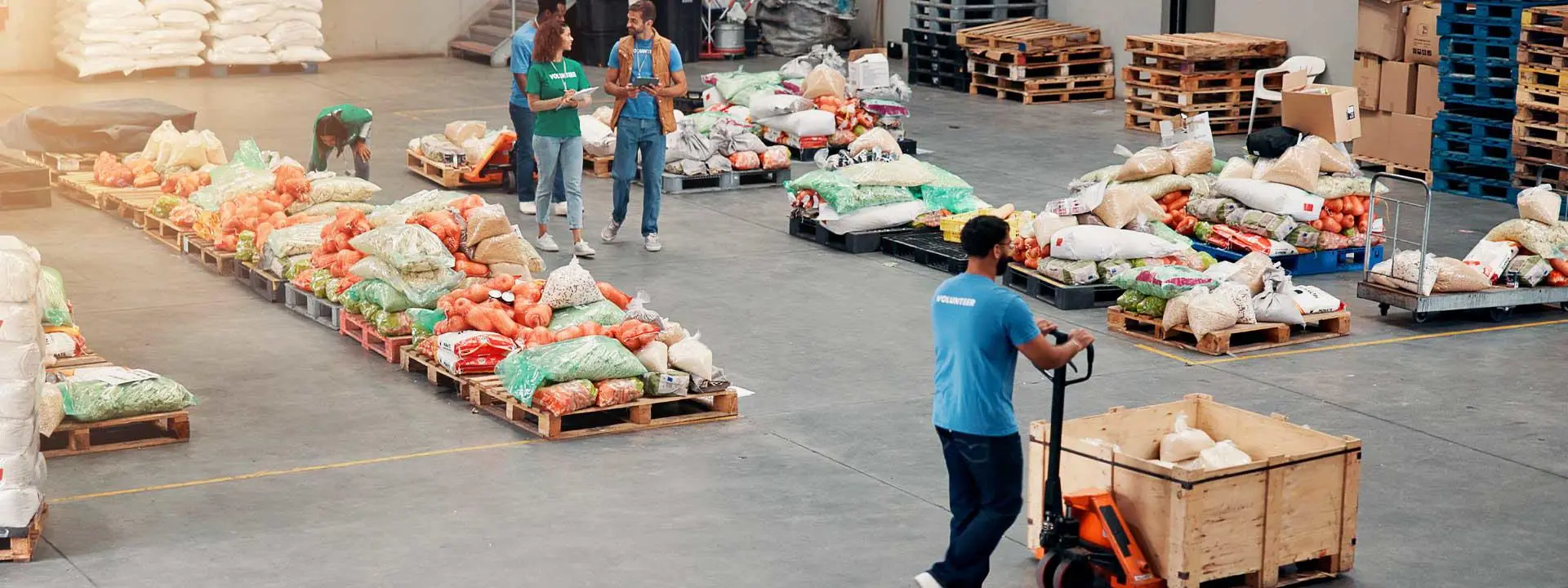 Volunteers organising and moving pallets of food supplies in a large warehouse with bags of vegetables and grains.