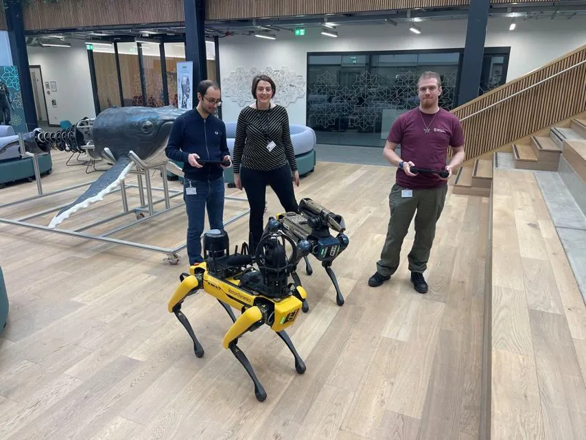 Research technicians from UKRAS-STEPs on secondment at the National Robotarium, alongside Project Manager Catherine Ciarka-White A group of three people stands in a modern indoor space with wooden flooring, open staircases, and contemporary furniture. Two of them are holding controllers while interacting with a yellow Boston Dynamics Spot robot, equipped with additional sensors and cameras.