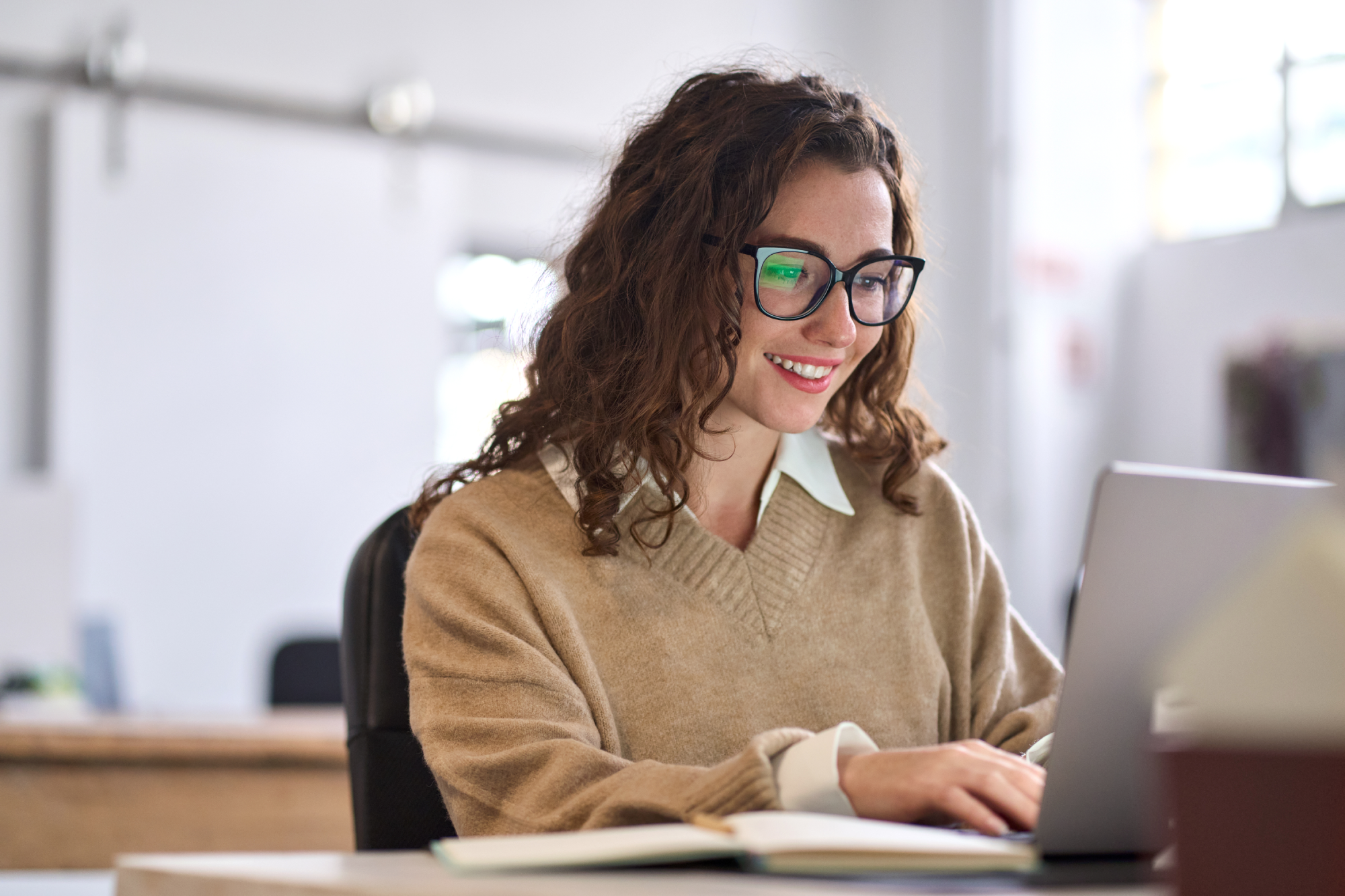 A woman wearing glasses smiles as she works on a laptop